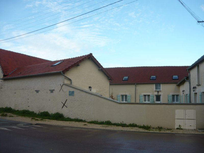 Vue depuis la rue après travaux : mur de clôture enduit beige crème, toitures neuves à velux, volets vert d'eau. Croix de Saint-André métalliques conservées sur le mur — détail authentique préservé.