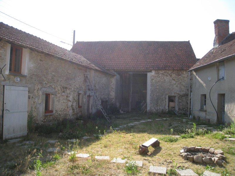 La cour intérieure avant travaux : grande grange à porte en bois, murs de pierre dégradés, mauvaises herbes, banc rustique.