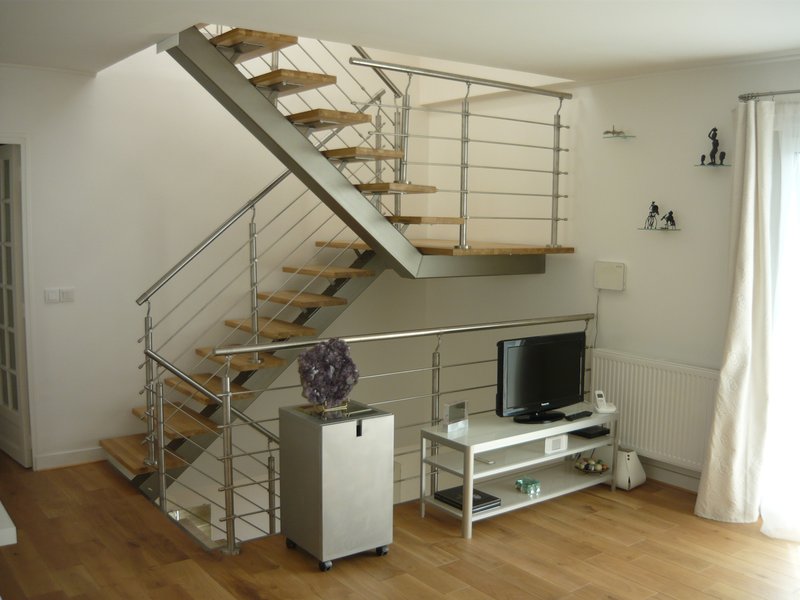 L'escalier dans son contexte de vie : volée à mi-hauteur, séjour en contrebas (parquet chêne, meuble TV), rideaux clairs.